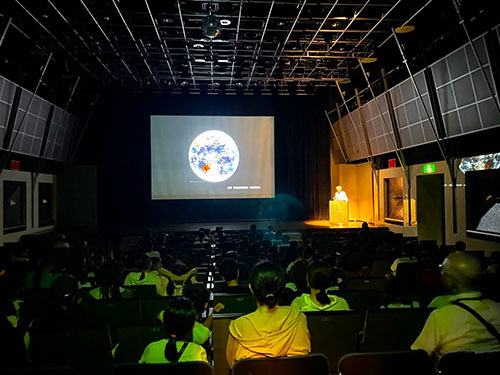 写真：当日の様子　正面スクリーンをたくさんの親子が椅子に座ってみている。