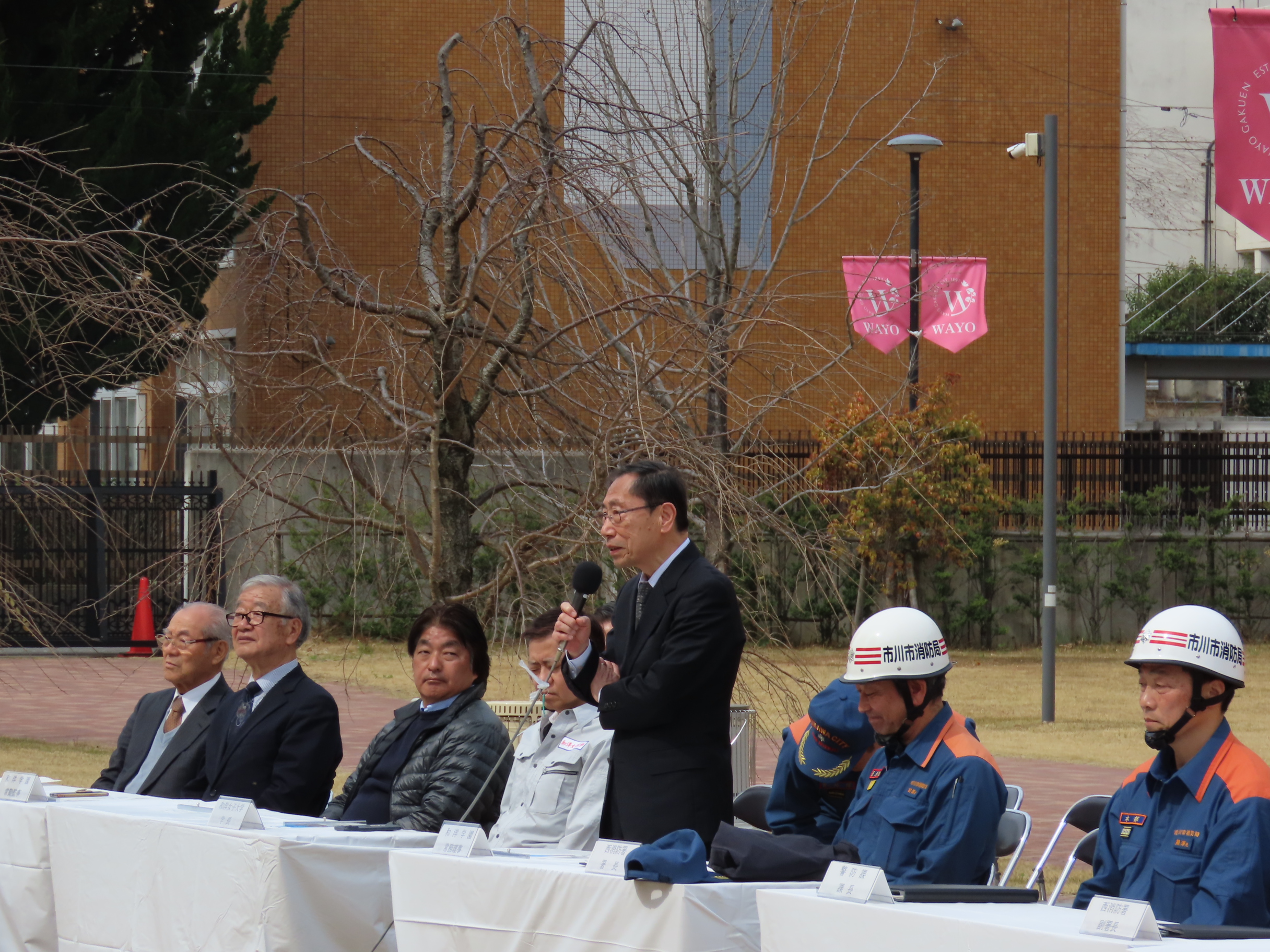 写真：立って挨拶する学長と、座る関係者たち