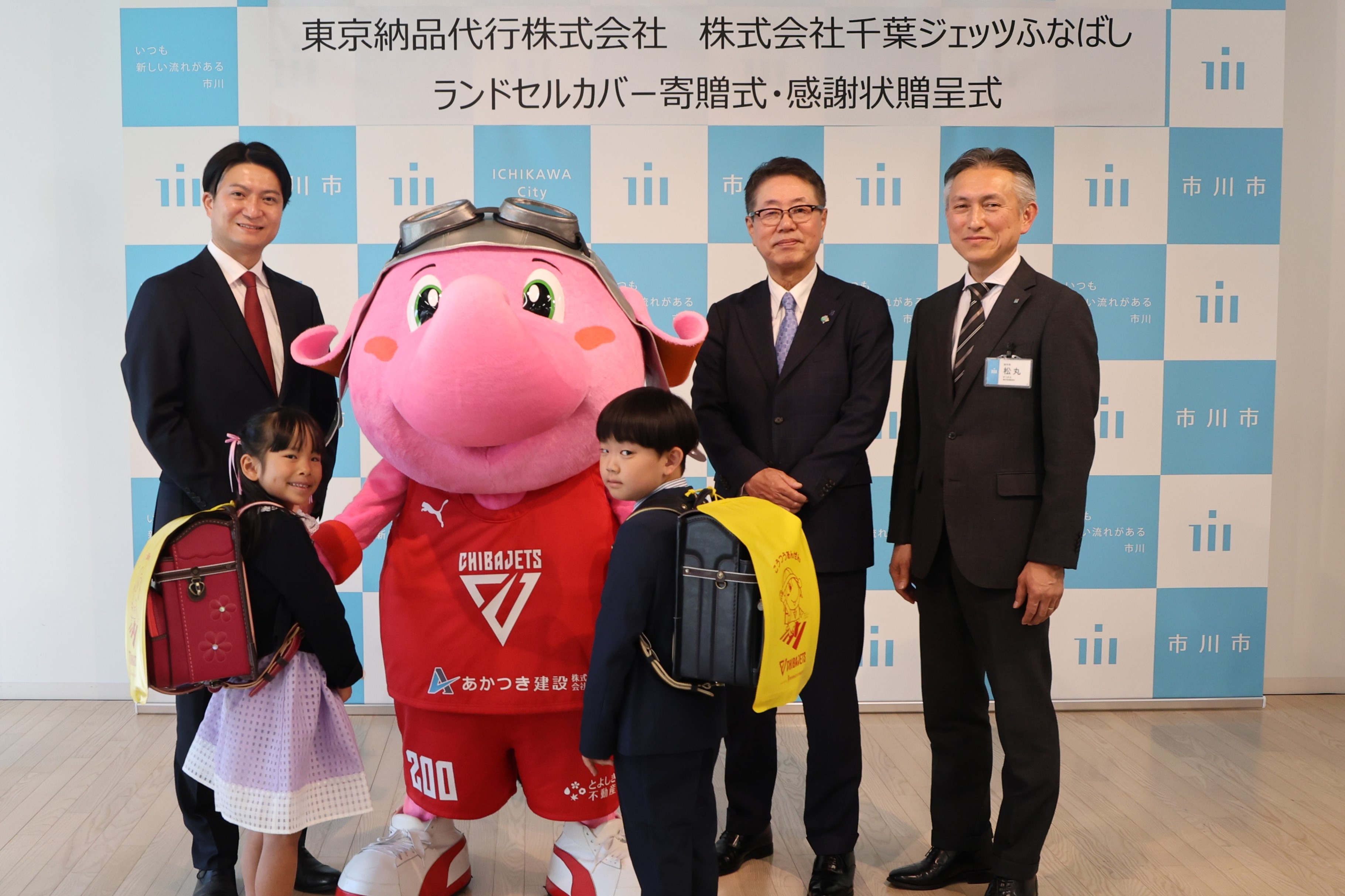 写真:寄贈されたカバーをのついたランドセルを背負った新入生代表のお二人と、田村代表取締役社長、ジャンボくん、大草代表取締役社長、松丸副市長