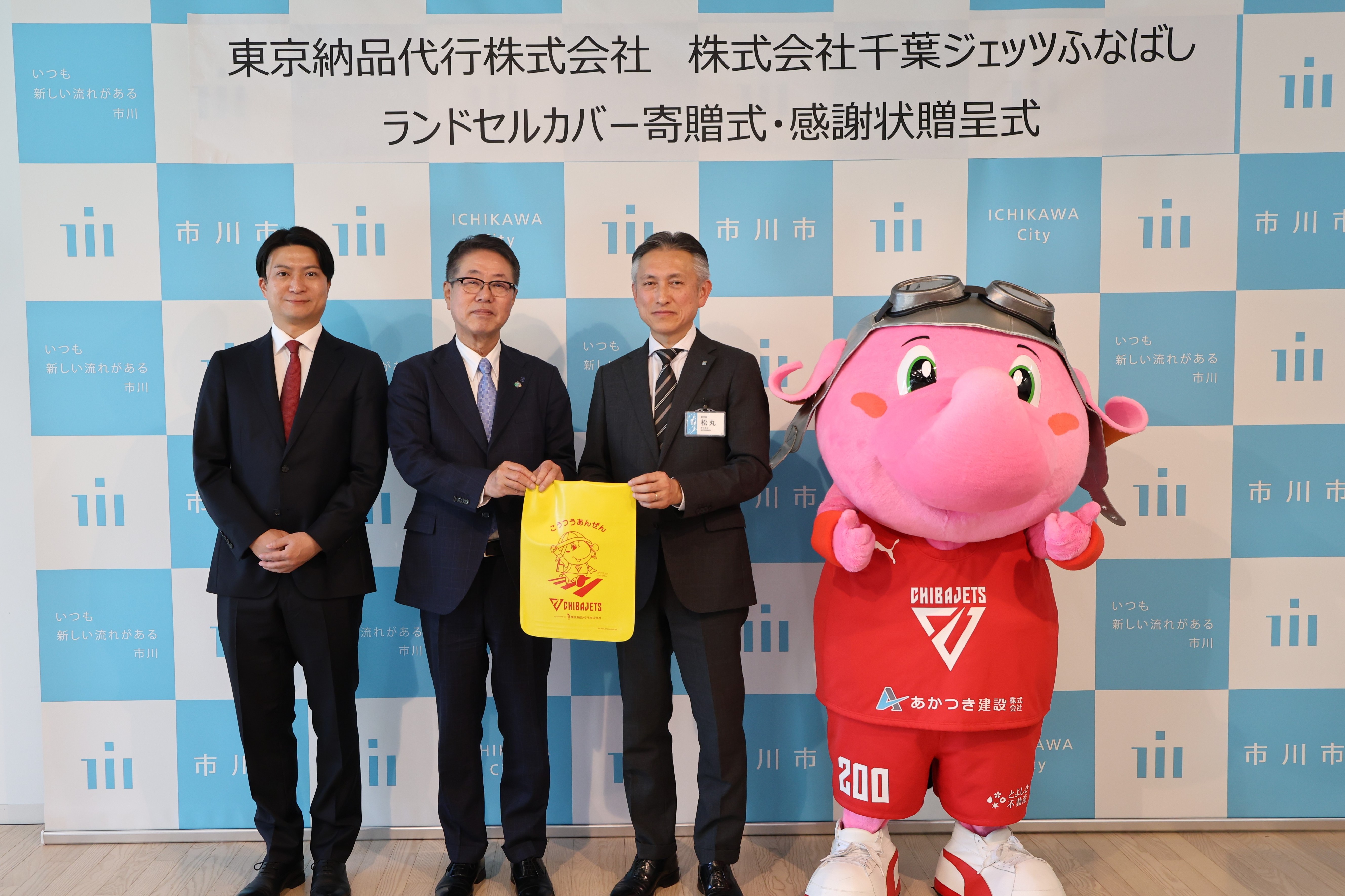 写真:田村代表取締役社長、ランドセルカバーを持った大草代表取締役社長と松丸副市長、ジャンボくんの集合写真
