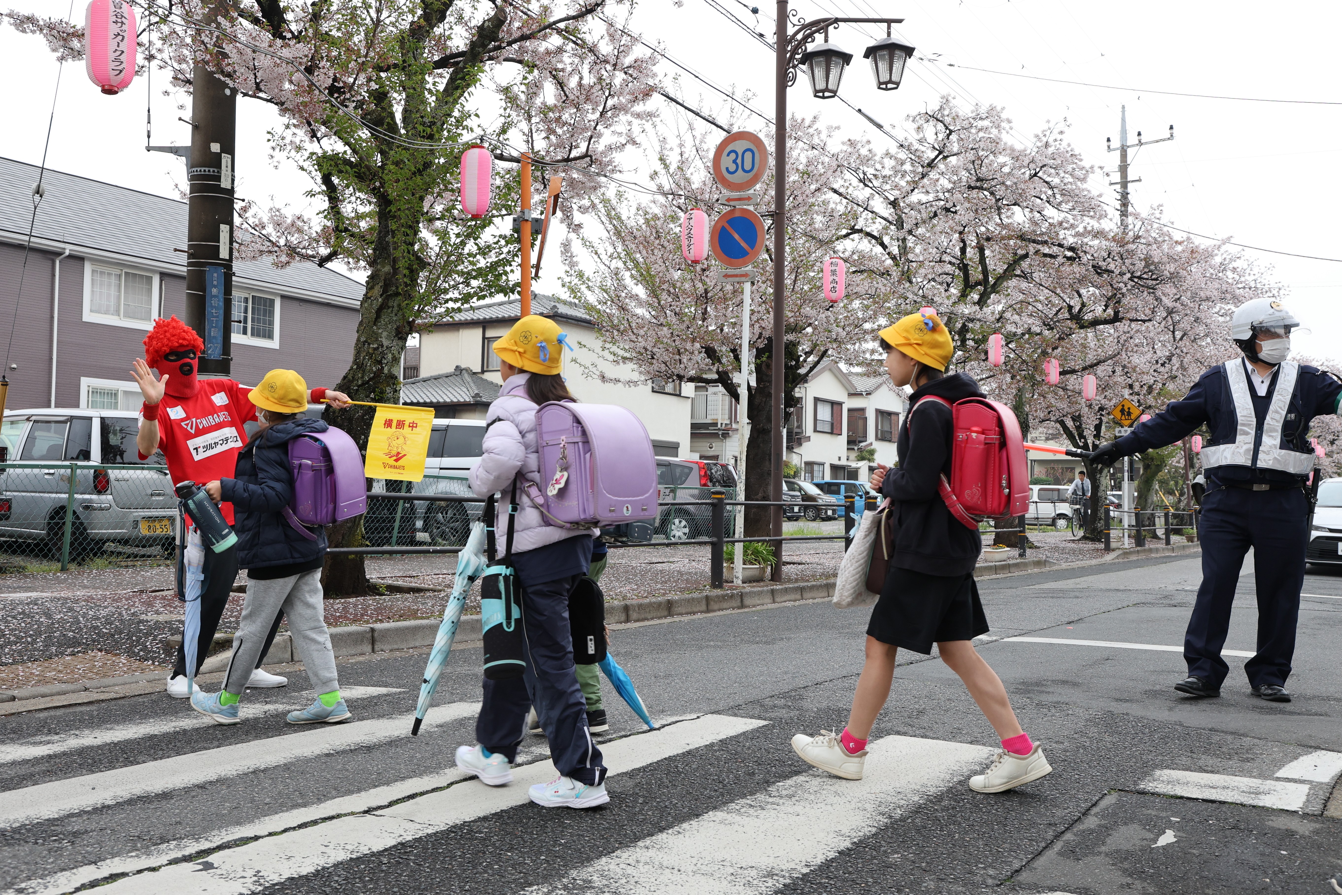 写真：登校中の児童と、交通安全旗を持って見守るマスク・ド・オッチー